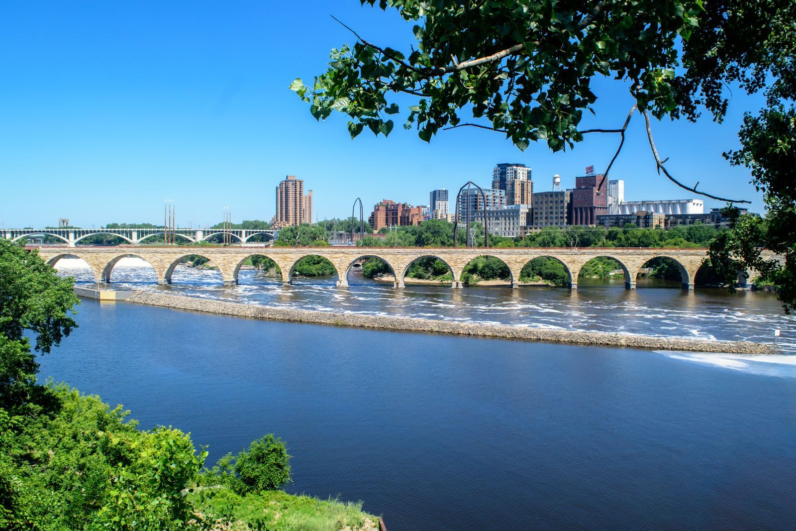 The Stone Arch Bridge crossing the Mississippi River in Minneapolis, with riverfront apartment towers and mill buildings behind it on a sunny day