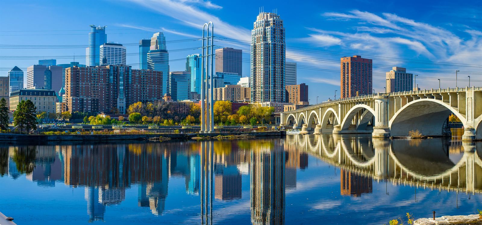 The Minneapolis skyline reflected in the Mississippi River, with the arched Third Avenue Bridge on a sunny autumn day