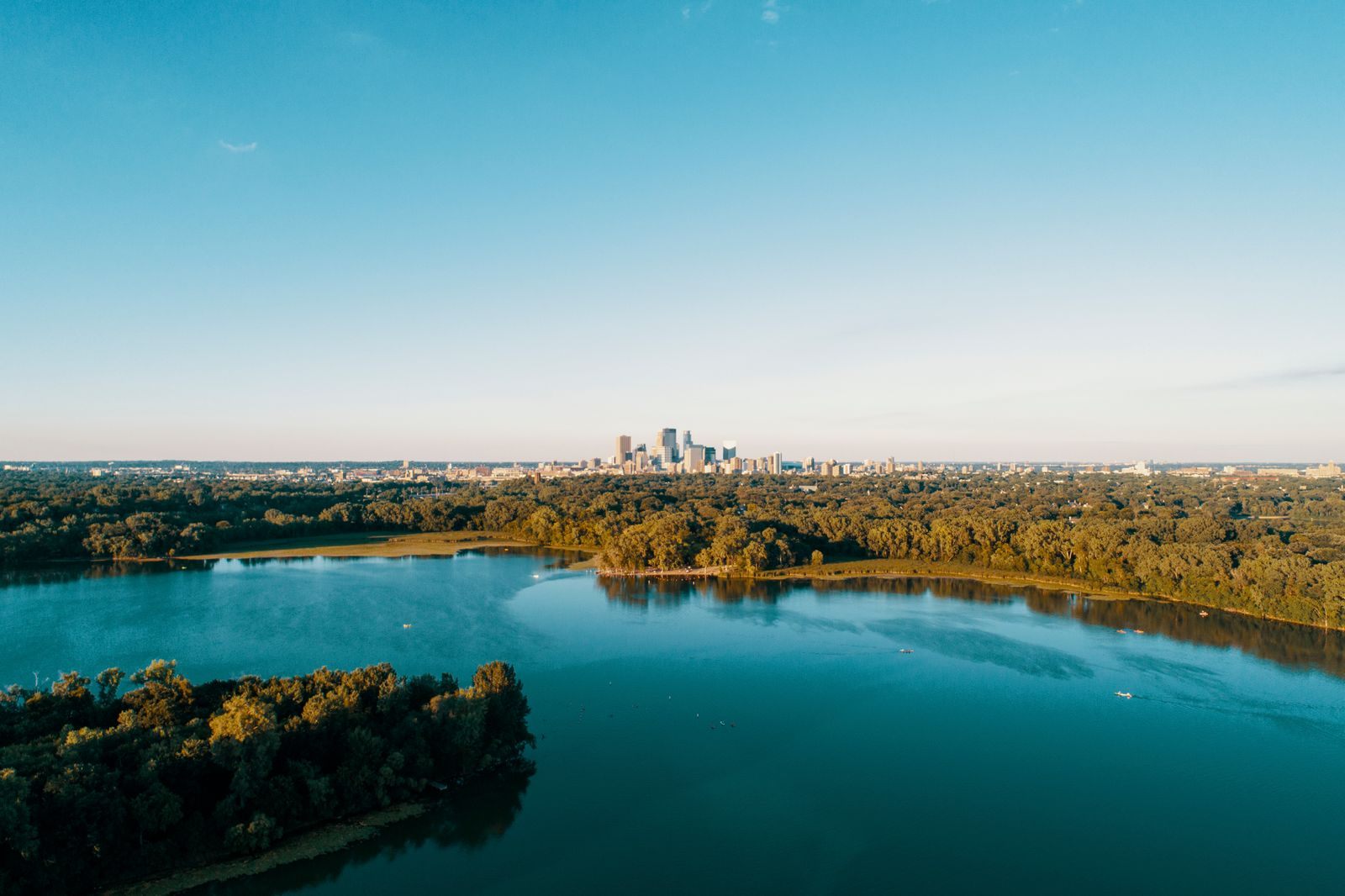 Aerial view of a tree-lined Minneapolis lake with small boats on the water and the downtown skyline on the horizon